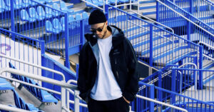 Loud Man wearing a black beanie, sunglasses, white t‑shirt and dark jacket standing on blue stadium bleachers.