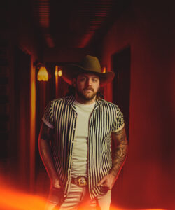 Matt Lang - Man with a cowboy hat stands in a dim, red-lit hallway wearing a striped shirt and white tee, looking at the camera with serious expression.