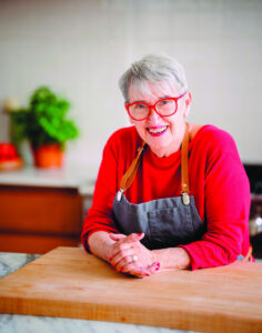 Portrait d’Elena Faita dans sa cuisine, souriante, assise devant une table de travail culinaire.