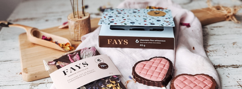 Pink heart-shaped chocolates on a dark chocolate shell, with FAYS boxes and a white cloth on a rustic wooden surface.