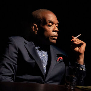 Portrait of a man in a navy suit smoking a cigarette in a dimly lit setting.