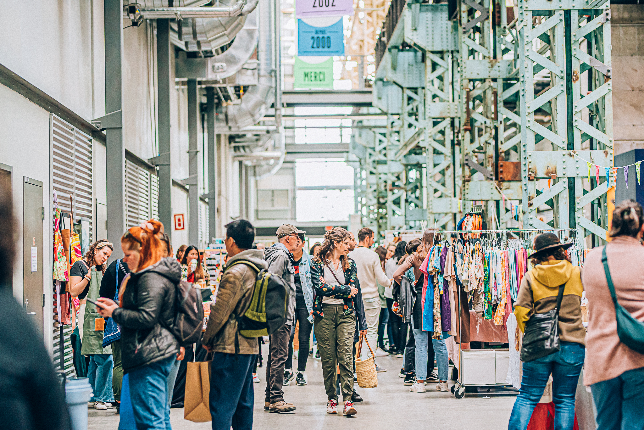 Busy indoor market aisle with shoppers browsing colorful clothing racks beneath green metal scaffolding and banners overhead.