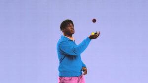 Young man in a bright blue hoodie juggling colorful balls against a purple background.