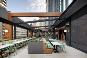Rooftop cafe with green chairs, marble-topped tables, and wooden planters under a metal and glass canopy in an urban setting
