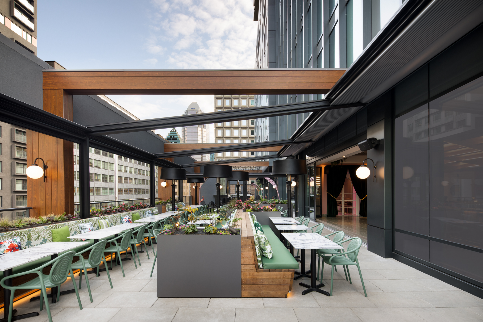 Rooftop cafe with green chairs, marble-topped tables, and wooden planters under a metal and glass canopy in an urban setting