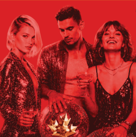Three people in sequined outfits posing at a party, with a gold gift bow visible in the foreground.