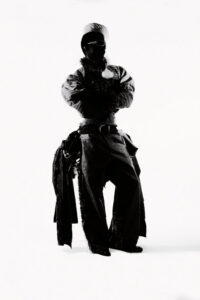 Silhouette of a person in an oversized coat and wide pants, arms crossed, wearing a headwrap and sunglasses against a white studio background.