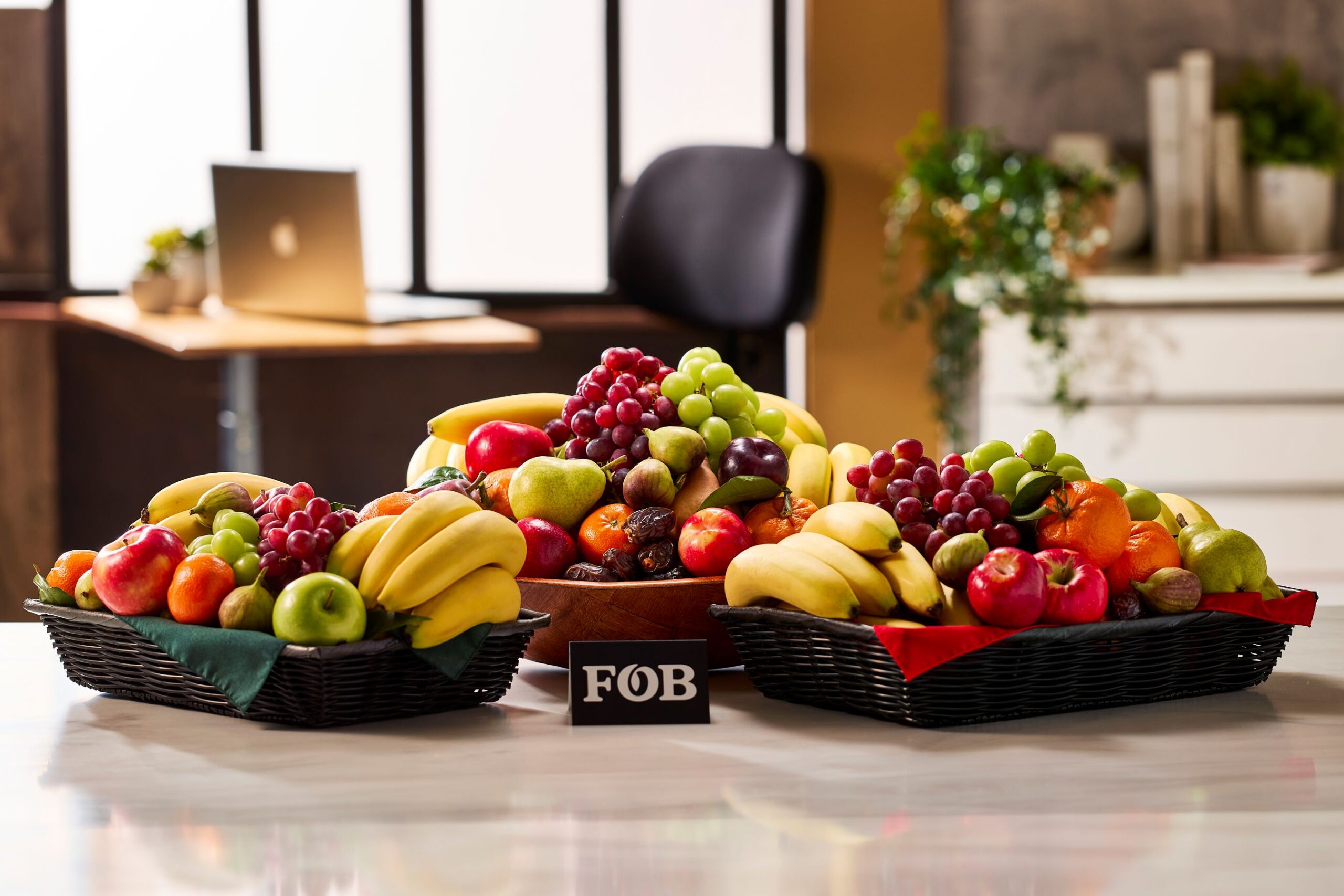 Three baskets of mixed fruit—bananas, grapes, apples, and citrus—on a bright showroom table with an FOB sign in front.