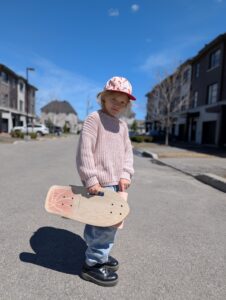 Un enfant souriant qui porte un casque de sécurité Coco Village et tient un skateboard en bois à l'extérieur.