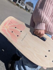 Close-up of a hand gripping a wooden longboard with a pink flower graphic and small hardware.