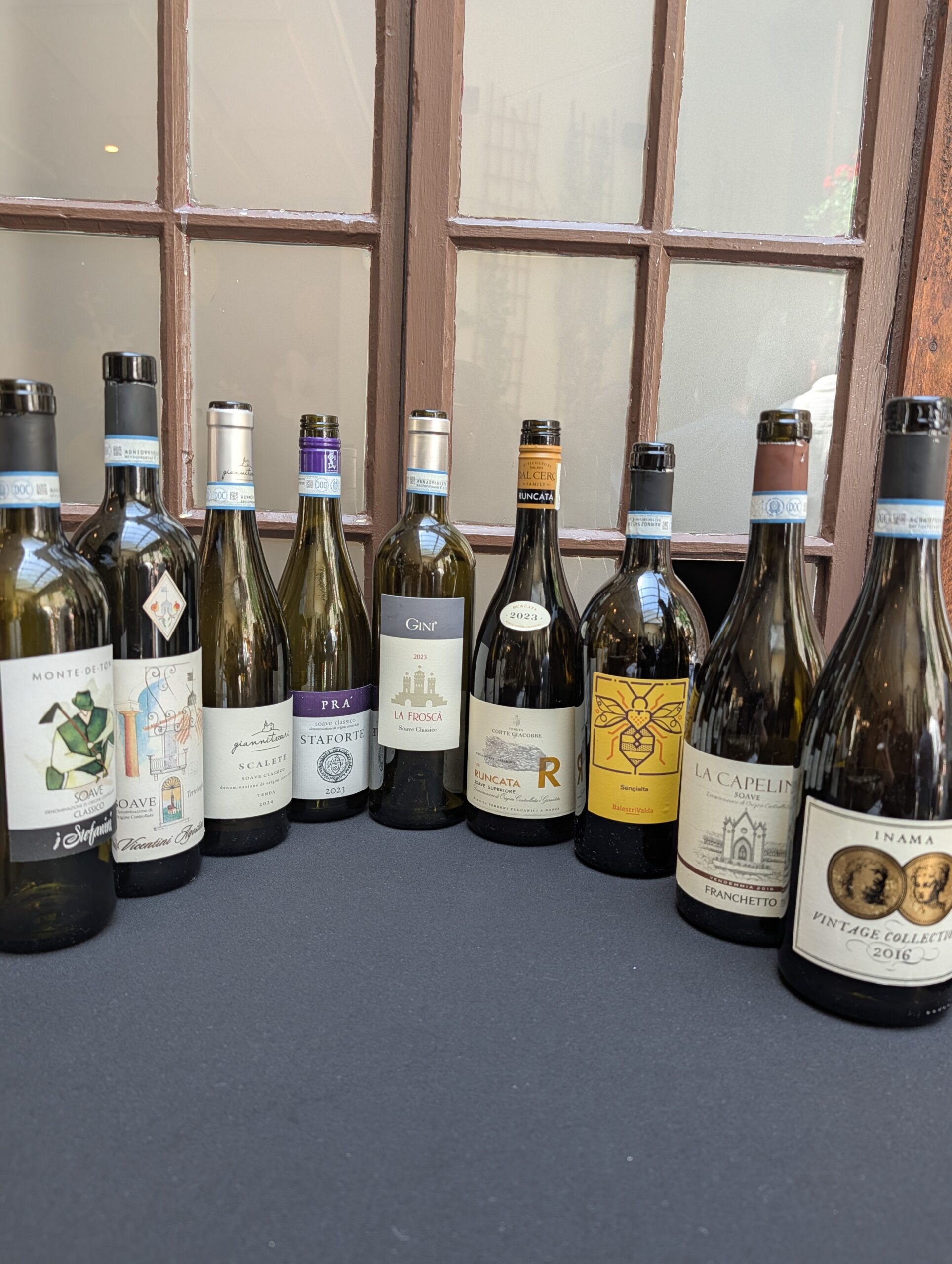 Vins de Soave - Row of wine bottles lined up on a dark surface with a wooden-framed window in the background