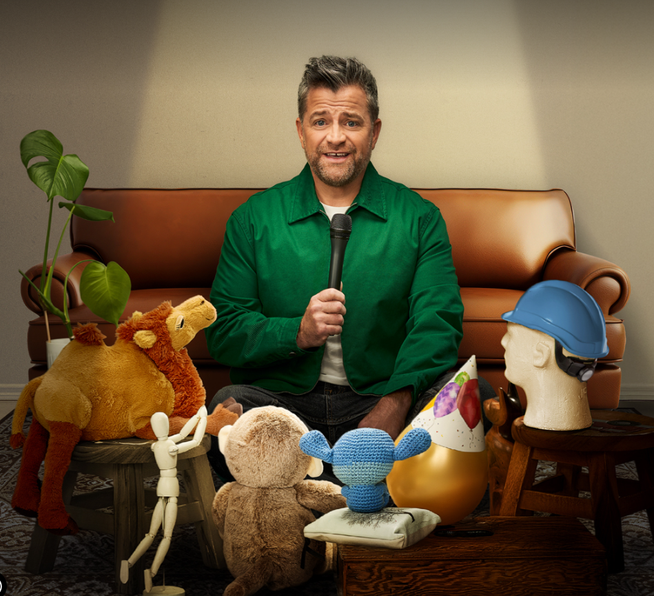 Patrick Groulx with a microphone sits on the floor among plush toys and a blue hard hat on a mannequin head, as if hosting a kids' show.