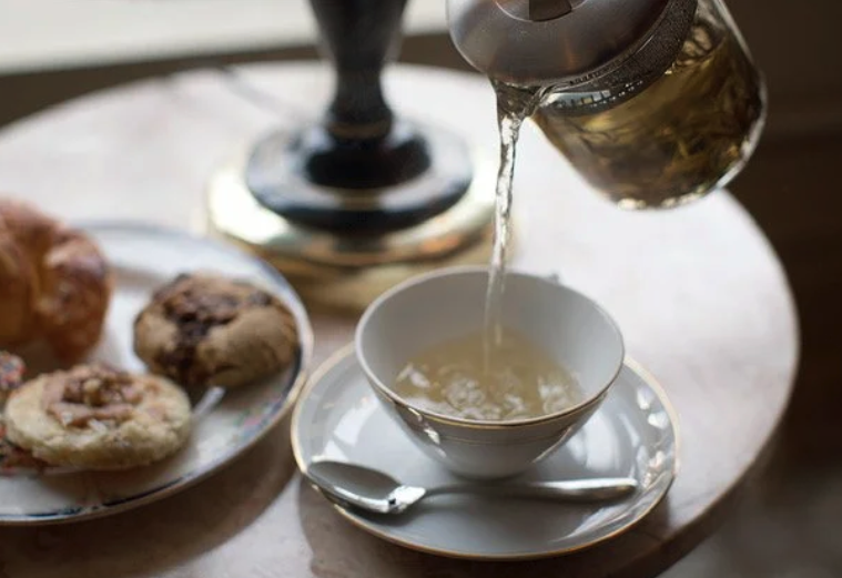 Hot water pours from a teapot into a white teacup on a saucer, with a plate of cookies nearby on a marble table.