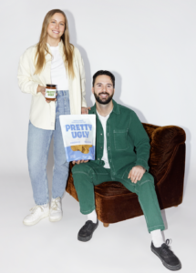 Smiling couple in a studio: woman stands with a jar labeled 'Pretty Ugly' while man sits on a brown chair holding a bag of Pretty Ugly tortilla chips.