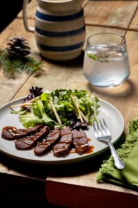Sliced beef with sauce on a white plate beside a mixed green salad and a fork on a wooden table.