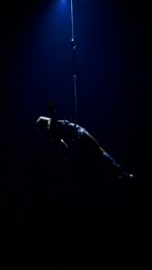 Acrobat suspended on a rope in a dark blue-lit scene, with one arm reaching upward.