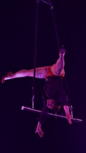 Two aerialists hang from a suspended rig in purple-lit stage, one upside down with red costume.