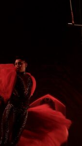Male dancer in a glittering black costume with a red cape, performing on a dimly lit stage under red lights.