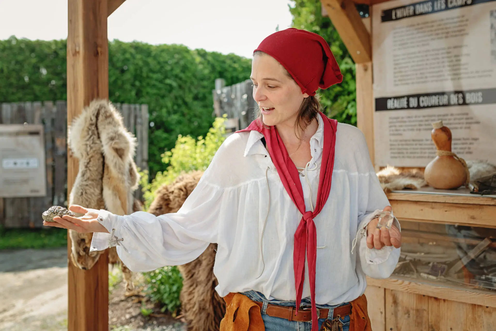 Woman in a white peasant blouse and red headscarf presenting a stone in her outstretched hand at an outdoor historical exhibit.