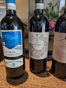 Three Michele Chiarlo wine bottles on a wooden surface: left bottle labeled Le Orme Barbera d'Asti with a blue artwork, middle bottle Palàs Barbaresco D.O.C.G. with circular pattern labels, right bottle similar Palàs Barbaresco D.O.C.G. label