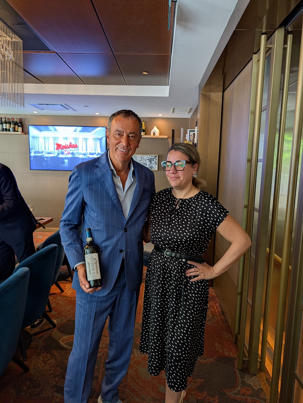 Two adults posing for a photo in a stylish lounge; the man in a blue pinstripe suit holds a wine bottle while the woman in a black polka-dot dress stands beside him, smiling.