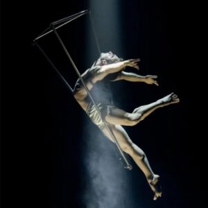 Male aerialist performing on an aerial hoop, suspended midair with arms and legs extended, lit by a spotlight against a dark background.