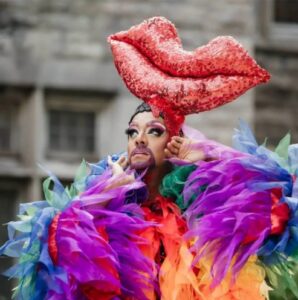 Drag performer wearing a huge red lipstick headpiece and a multicolored feathered costume performing outdoors.