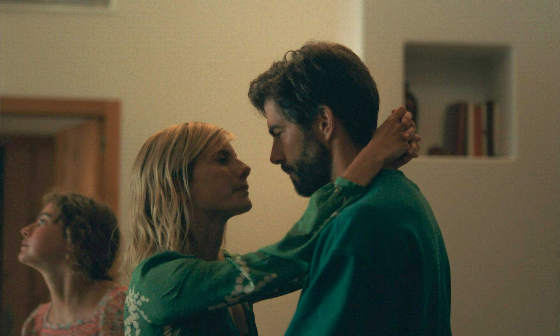 Un film - Qui brille au combat -Couple in green gowns embrace, gazing at each other in a softly lit room with a blurred figure in the background