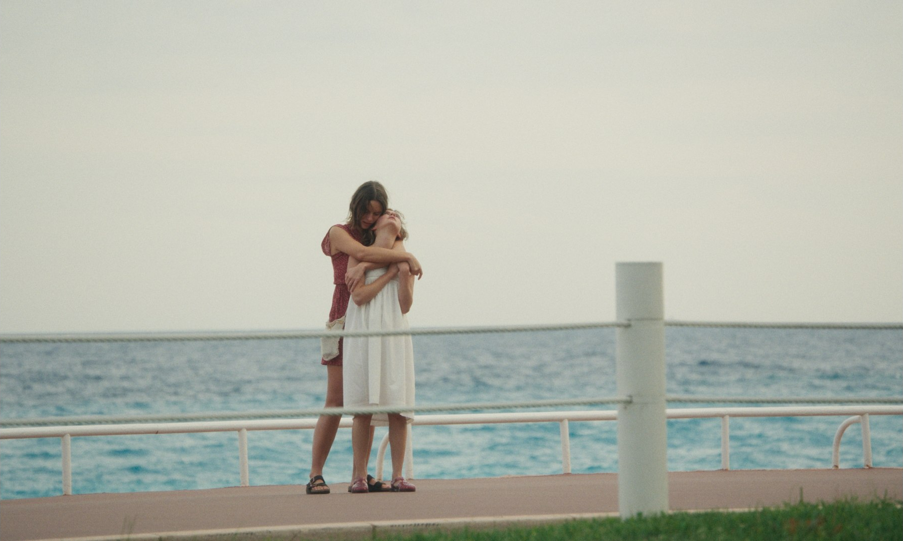Un film - Qui brille au combat -Two women embrace on a seaside pier, with blue ocean behind them and a pale sky above.