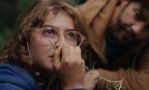 Un film - Qui brille au combat - Young woman with pink glasses sniffing a lavender sprig; a bearded man sits blurred in the background outdoors.