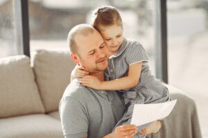 Father holding a young girl who hugs him while they read a letter together on a sofa, sunlit scene.