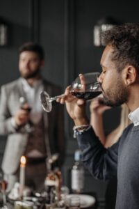 Close-up of a man drinking red wine from a glass at a dimly lit dinner party; another guest is blurred in the background with a candlelit table in view.