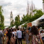 Crowd of people at an outdoor market with white vendor tents along a tree-lined path and a tall clock tower in the distance.