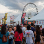 Crowd at a street food festival with white tents and a large Ferris wheel in the background.