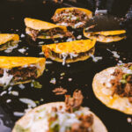 Tacos with beef, onions, and cheese cooking on a hot griddle, a gloved hand in the background.