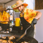 Chef in a yellow patterned shirt and straw hat grills shrimp on a large pan at an outdoor festival stall under a tent.