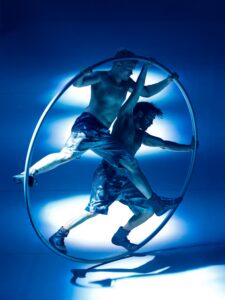 Two shirtless male acrobats perform on a large aerial hoop under vivid blue lighting, gripping the hoop and balancing mid-air by their arms and legs a circle high above the ground.