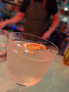 Close-up of a pale pink cocktail in a glass with a small orange slice garnish, blurred colorful bar scene in the background.