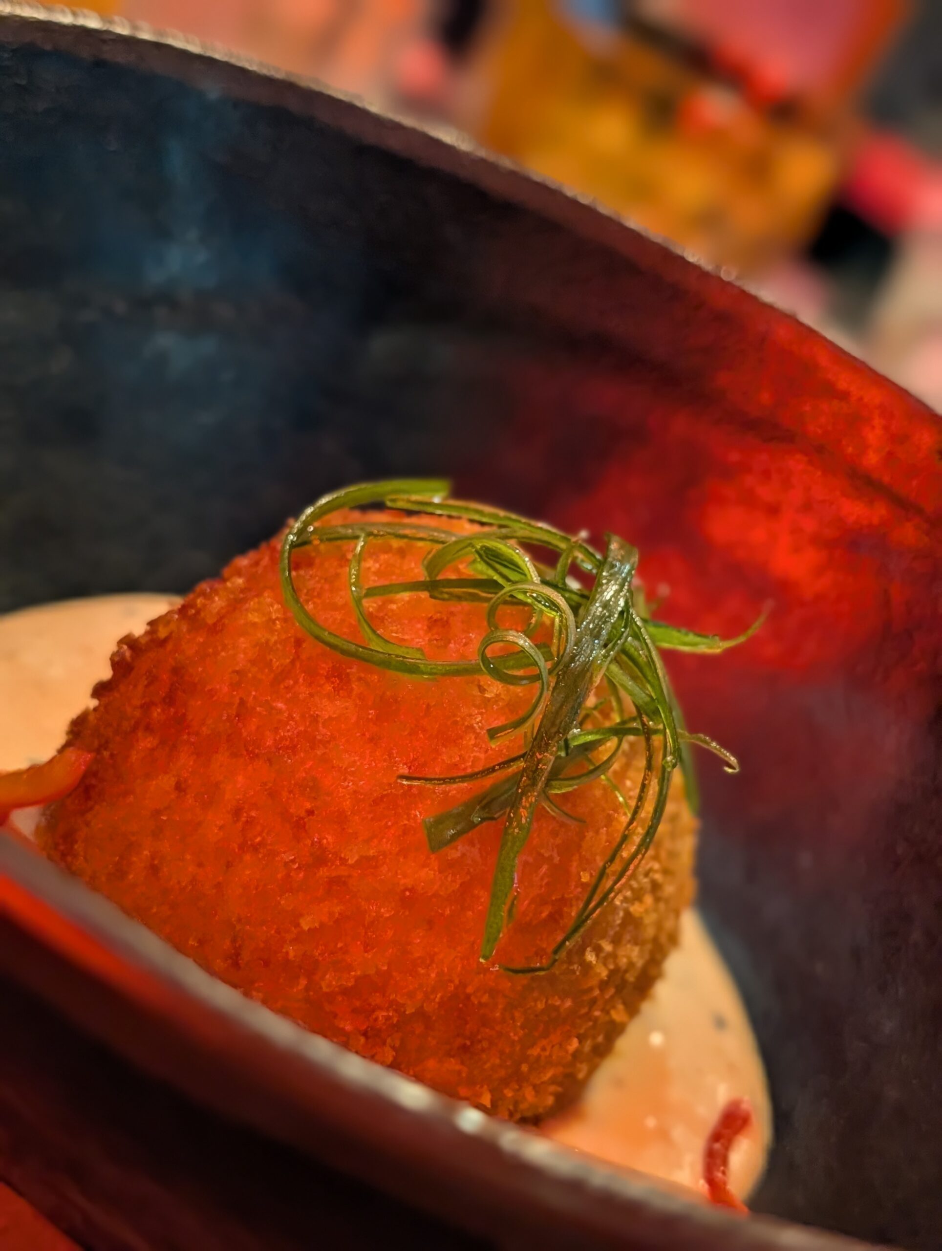 Close-up of a crispy orange croquette topped with curled green onion garnish on a plate. Tiradito