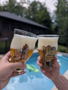 Two people toast with decorated beer glasses, foamy tops visible, poolside in the background.