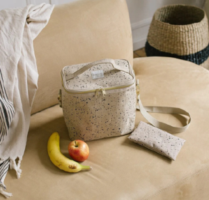 Beige speckled insulated lunch bag with shoulder strap and matching pouch on a tan sofa, next to a banana and an apple