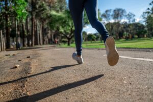 Close-up of a person running/jogging on a park path, blue leggings and white sneakers in mid-stride with trees in the background.