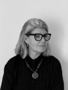 Portrait of a middle-aged woman with gray hair and bold black glasses, wearing a black shirt and a large circular pendant necklace, looking to the side with a neutral expression.